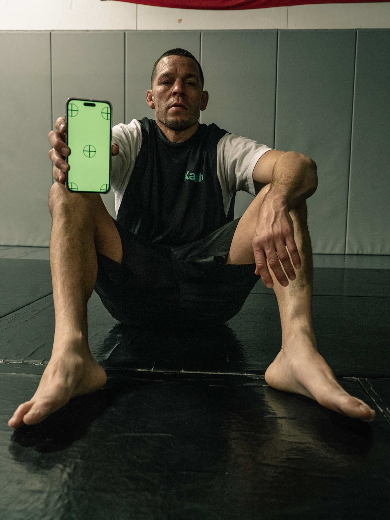 Nate Diaz seated on the mats holding a phone toward the camera